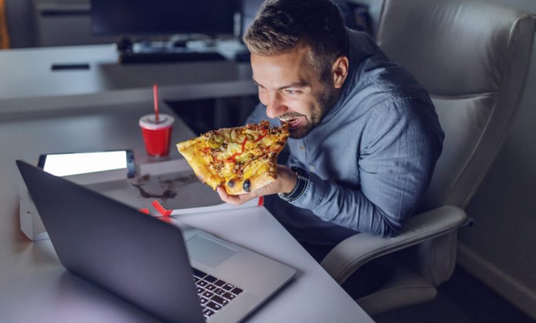 Young Caucasian architect eating pizza at office. On desk laptop. Late night work concept.